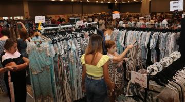 peças de roupas penduradas em araras e mulheres olhando e mexendo. Ambiente é de um grande bazar. #paratodosverem