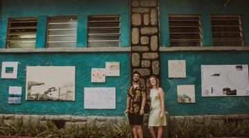 um homem e uma mulher estão à frentne de quadros expostos no muro da fachada do aquário. #paratodosverem 