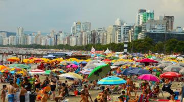 praia lotada em dia de sol com muitos guarda-sóis armados. #paratodosverem 