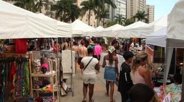barracas com produtos em exposição em uma praça a céu aberto com pessoas circulando. #paratodosverem 