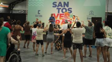 pessoas dança sob tenda, de costas para a foto e de frente para um palco onde um grupo se apresenta. #paratodosverem 