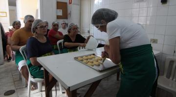 mulher está moldando pães sobre uma forma. Alunas ao fundo assistem. #paratodosverem