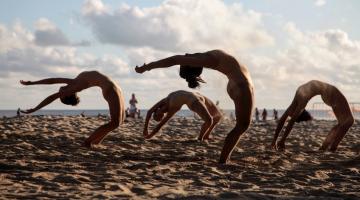 bailarinas dançam na areia e tem o tronco curvado para trás em busca de atingir o chão com as mãos. #paratodosverem