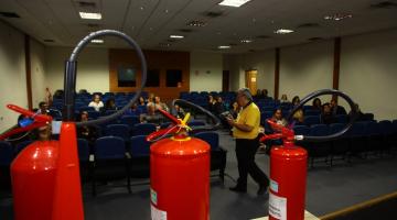 Instrutor conversa com público em auditório com três extintores de incêndio em primeiro plano. #pratodosverem