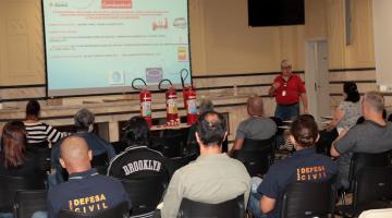 servidores estão sentados em auditório, de costas para foto, assistindo palestra. Há um telão na parede com textos sobre incêndio. Sobre uma mesa estão três extintores . #paratodosverem