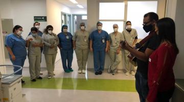 #pracegover Homem com violão e mulher se apresentam perante nove profissionais de saúde em corredor hospitalar