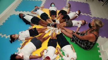 Crianças deitadas em círculo, juntas com professora, com todos encostando os pés. #pratodosverem