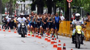 Atletas estão correndo em faixa de pista delimitada por cones. À frente e ao lado deles, dois motociclistas batedores. #Pracegover