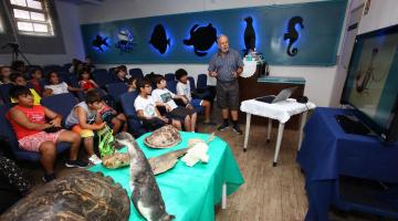 Mesa com animais marinhos taxidermizados em primeiro plano. Ao fundo, crianças estão sentadas no auditório atentas ao que um homem em pé está falando. #Pracegover