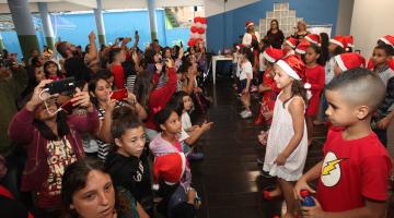 Crianças e adultos assistem apresentação de outras crianças que estão usando capuz de papai noel. #Pracegover