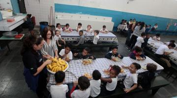 Alunos estão no refeitório tomando lanche. Todos estão sentados em torno de mesas. #Pracegover