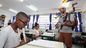 músico está em pé, com guarda chuva nas costas, tocando com aluno que está sentando tocando flauta #pracegover 