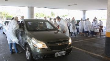 Veículo está parado em estacionamento. Pessoas vestidas aventais brancos, máscaras e luvas estão em volta. #Paratodosverem