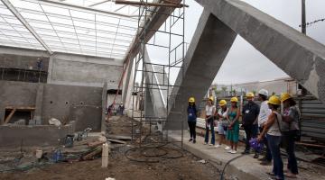 Pessoas estão em pé e paradas em uma obra. Elas estão abaixo de uma grande estrutura de concreto