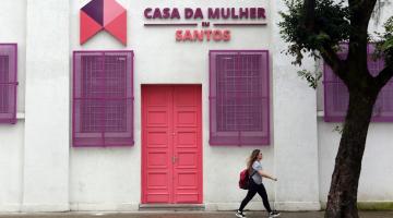 fachada da casa da mulher com uma mulher passando à frente, na calçada. Há também o troco de uma grande árvore. #paratodosverem 