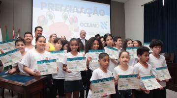 crianças posam para foto segurando certificados. Ao fundo há um telão onde se lê premiação. #paratodosverem