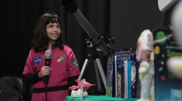 menina está segurando microfone ao lado de mesa com telecópio e outros objetos que tem ligação com o espaço. #paratodosverem