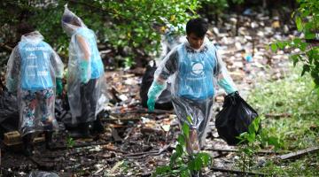 JOvens usando luvas estão em mangue recolhendo residuos e depositando em sacos. #paratodosverem