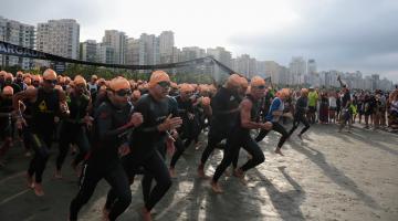 atletas uniformizados com roupa de natação e toca correm na praia em direção ao mar para prova de nado. #paratodosverem 