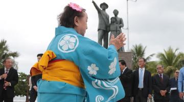 Mulher vestida com roupa típica dança em frente a monumento em homenagem à imigração, com estátuas de três pessoas formando uma família. #paratodosverem