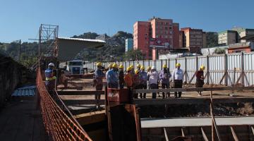 Obras na entrada da Cidade, com conjunto habitacional ao fundo. Pessoas circulam pela obra. #Pracegover