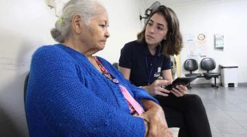 Estagiária da Ouvidoria conversa com idosa usuária de policlínica. Ambas estão sentadas. A jovem faz anotação em um tablet