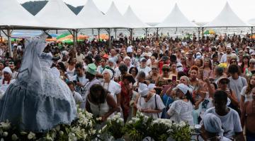 imagem da santa de costas em primeiro plano à esquerda e multidão em frente. Há tendas armadas na lateral do ambiente a céu aberto. #paratodosverem 