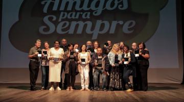 servidores reunidos no palco segurando medalhas posam para foto. Ao fundo, na parede se lê amigos para sempre. #paratodosverem