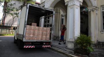 caminhão cheio de cestas básicas com a porta de trás aberta. Ao fundo, um homem carrega uma caixa para dentro de um prédio. #paratodosverem