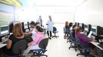 Sala de aula com diversos alunos diante de bancadas com computadores. #Pracegover