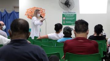 pessoas estão sentadas em sala de espera assistindo palestra feita por uma mulher. #paratodosverem