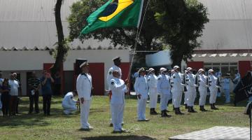 marinheiros uniformizados hasteando a bandeira do brasil ao ar livre sobre um gramado. #paratodosverem 