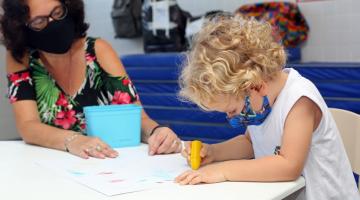 criança de máscara desenha observada por professora #paratodosverem 