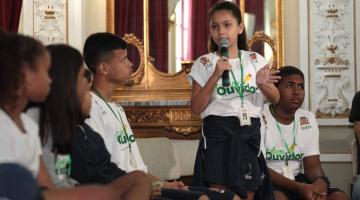 Alunos ouvidores estão sentados em cadeiras no salão nobre do paço. Uma menina está em pé e fala ao microfone. Todos usam camisetas alusivas ao programa. #Pracegover