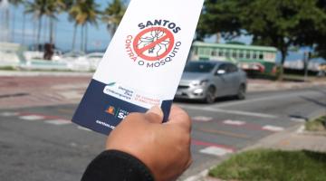 close sobre mão direita que segura folheto onde se lê: Santos contra o mosquito. A avenida da praia está ao fundo. #paratodosverem 
