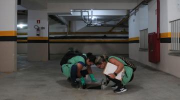 duas agentes uniformizadas agachadas examinam um ralo numa garagem de prédio. #paratodosverem 