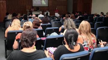 várias pessoas sentadas em um auditório e uma pessoa falando ao fundo à frente de um telão. #paratodosverem