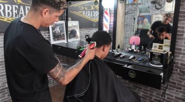barbeiro está em pé passando máquina em cabelo de homem que está sentado de frente para o espelho. #paratodosverem