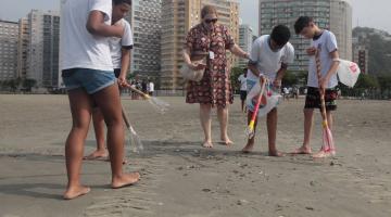 estudantes uniformizados andam na areia da praia catando lixo com uma garra feita com tubo pvc e garrafa pet. #paratodosverem 