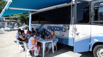 Ônibus do procon móvel está parado. Pessoas estão sentadas sendo atendidas por recepcionistas. #paratodosverem