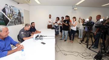 Três homens estão sentados a uma mesa. Sobre a mesa há microfones. Repórteres estão em pé diante dos homens que dão entrevista