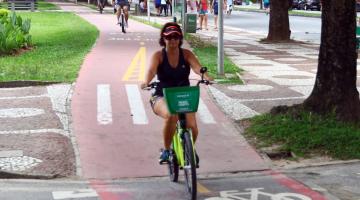 Mulher está pedalando bicicleta do bike santos em ciclovia. #Pracegover