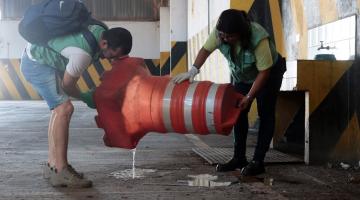 Dois agente seguram cone de trânsito para despejar água contida no interior. #paratodosverem 