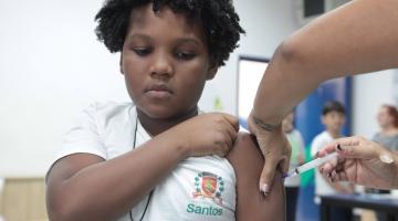 criança é vacinada no braço. Ao fundo, outras crianças olham. #paratodosverem 