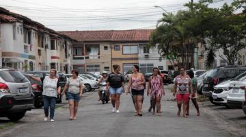 mulheres caminham por rua de uma vila. Nas laterais, os carros estão estacionados a 90 graus. #paratodosverem 