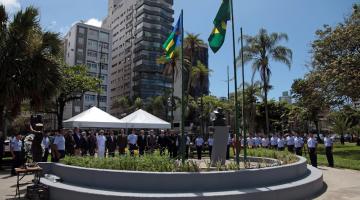 duas bandeiras hasteadas em canteiro no jardim da orla. Várias pessoas estão em pé sob tendas em cerimônia. #paratodosverem