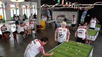 várias duplas jogando futebol de botão no Museu Pelé decorado por bandeiras de times participantes da copa. #paratodosverem