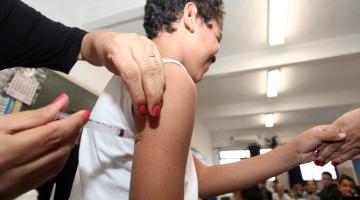 Mãos femininas aplicam injeção em braço de adolescente. O adolescente segura na mão de alguém. #Paratodosverem