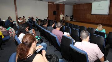 Pessoas estão sentadas em poltronas de auditório. Elas estão atentas a um homem que fala ao fundo. Na parede há uma imagem projetada em um telão. #Pracegover