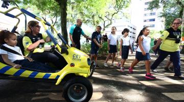 Minibugue aparece em primeiro plano com uma mulher pilotando e uma criança sentada ao lado. À direita, crianças atravessam na faixa. #Pracegover